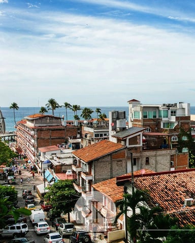 Coastal town with terracotta-roofed buildings, palm trees, ocean view, and modern white building on left