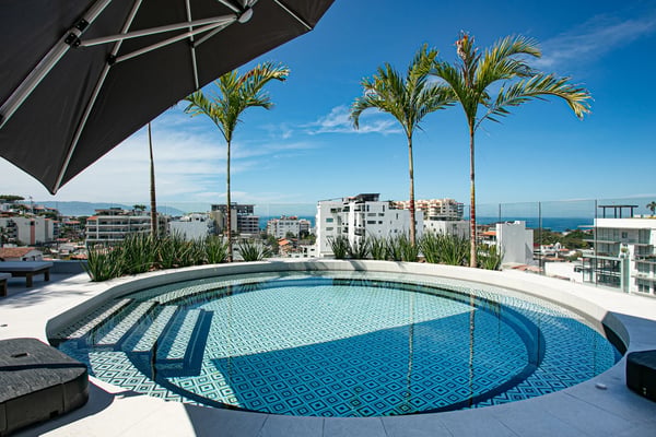 Rooftop pool with geometric tile pattern, palm trees, black umbrella, and ocean view beyond coastal buildings