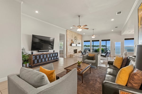 Open-plan living room with waterfront views, stone fireplace, ceiling fans, and multiple seating areas with yellow accents