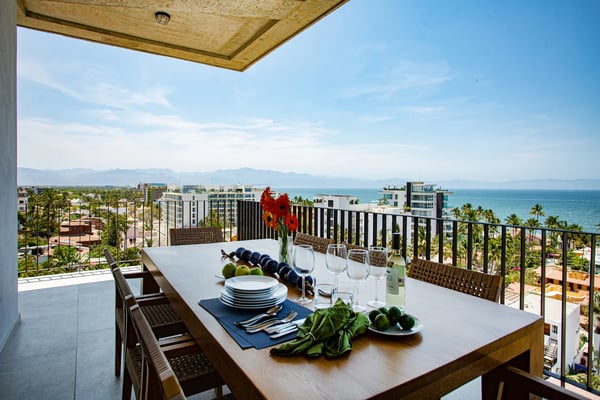 Beachfront balcony with dining table set for meal, ocean and mountain views, palm trees and coastal buildings below