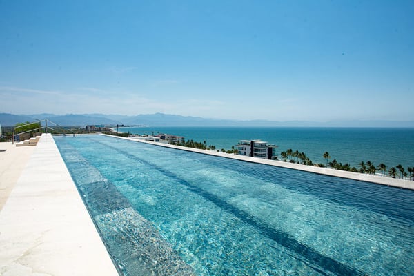 Rooftop infinity pool overlooking Mediterranean Sea with palm trees, coastal buildings, and mountains in distance