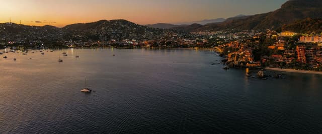 Aerial view of a Mexican bay at golden-hour sunset with city lights and mountains in the distance