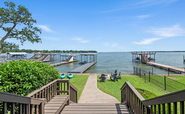 Waterfront property with wooden deck, lawn, boat docks, and calm lake view under blue sky