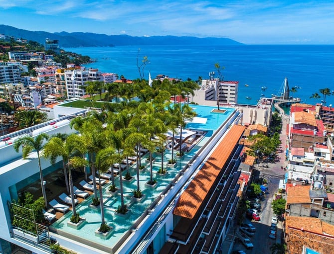 Aerial view of beachfront resort with turquoise pools, palm trees, and blue ocean with mountains beyond